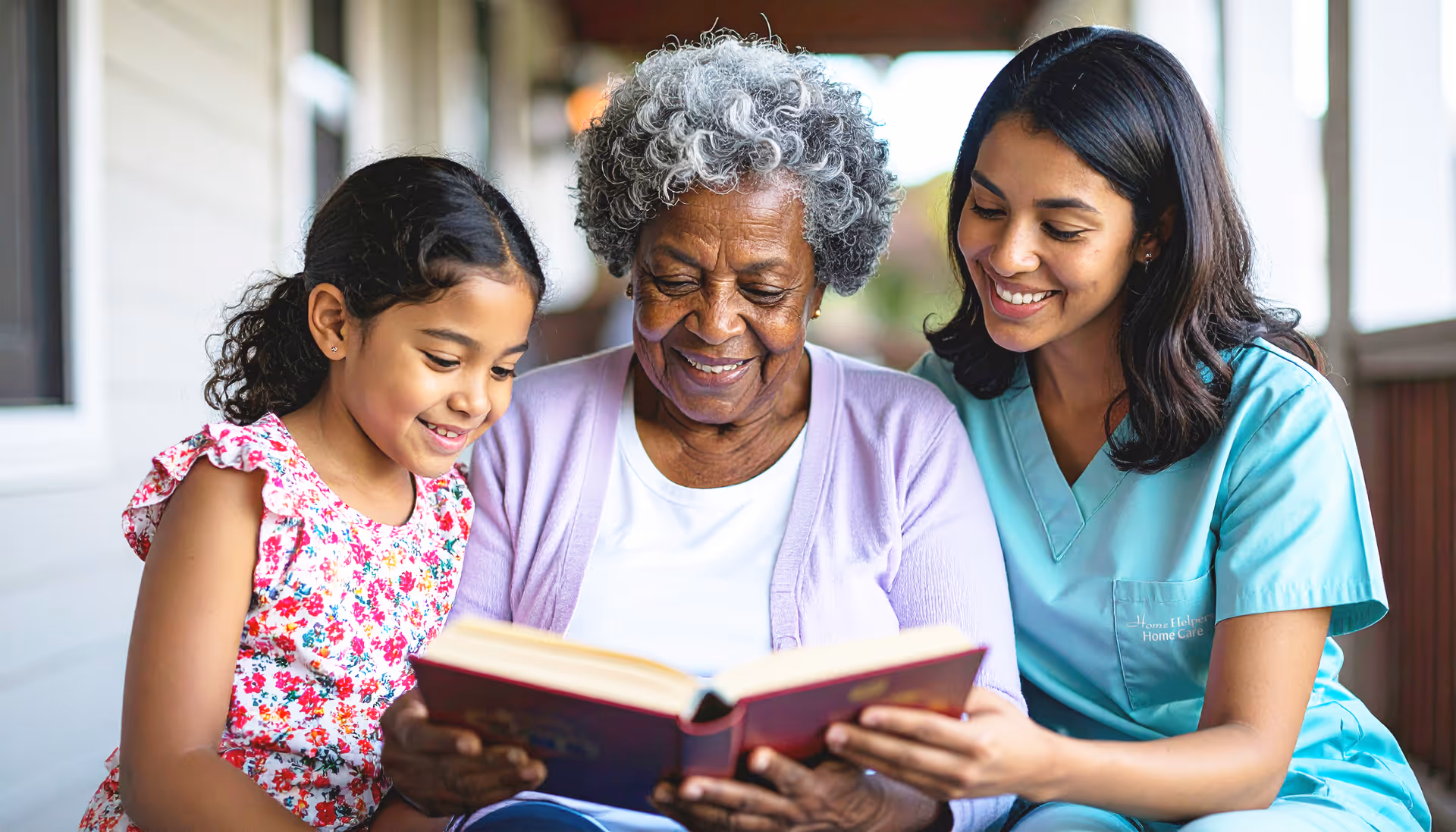Elderly woman, child, and caregiver read a book together on a porch.