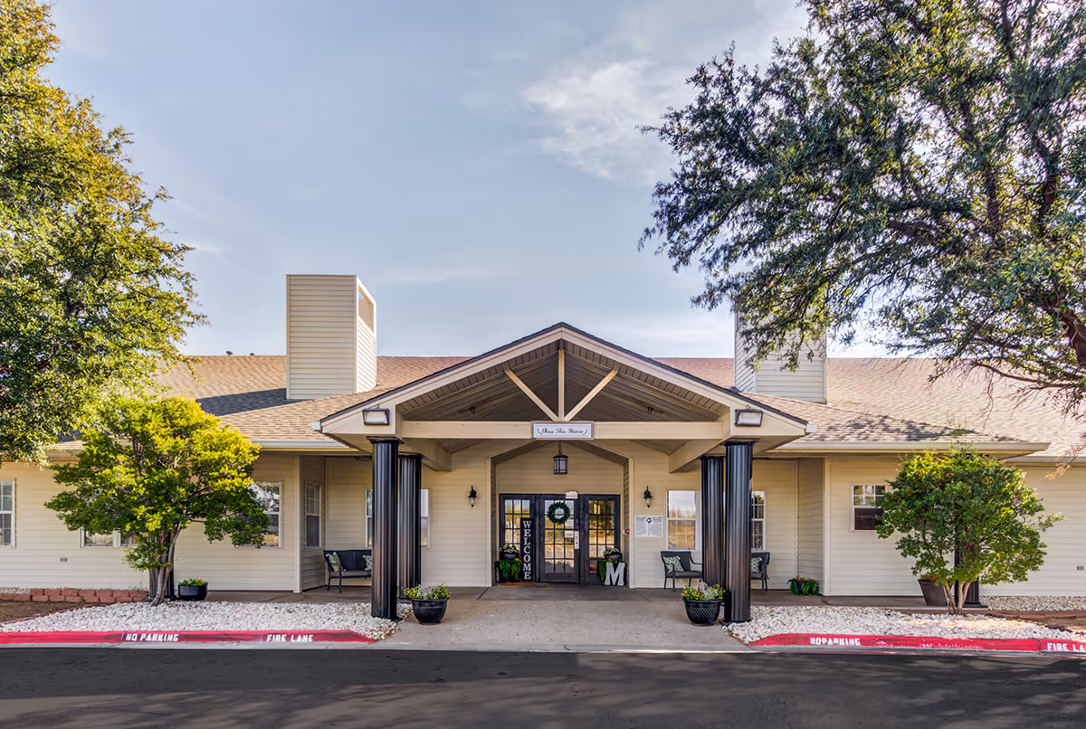 Front of a senior living facility with a covered entrance, trees, and seating.