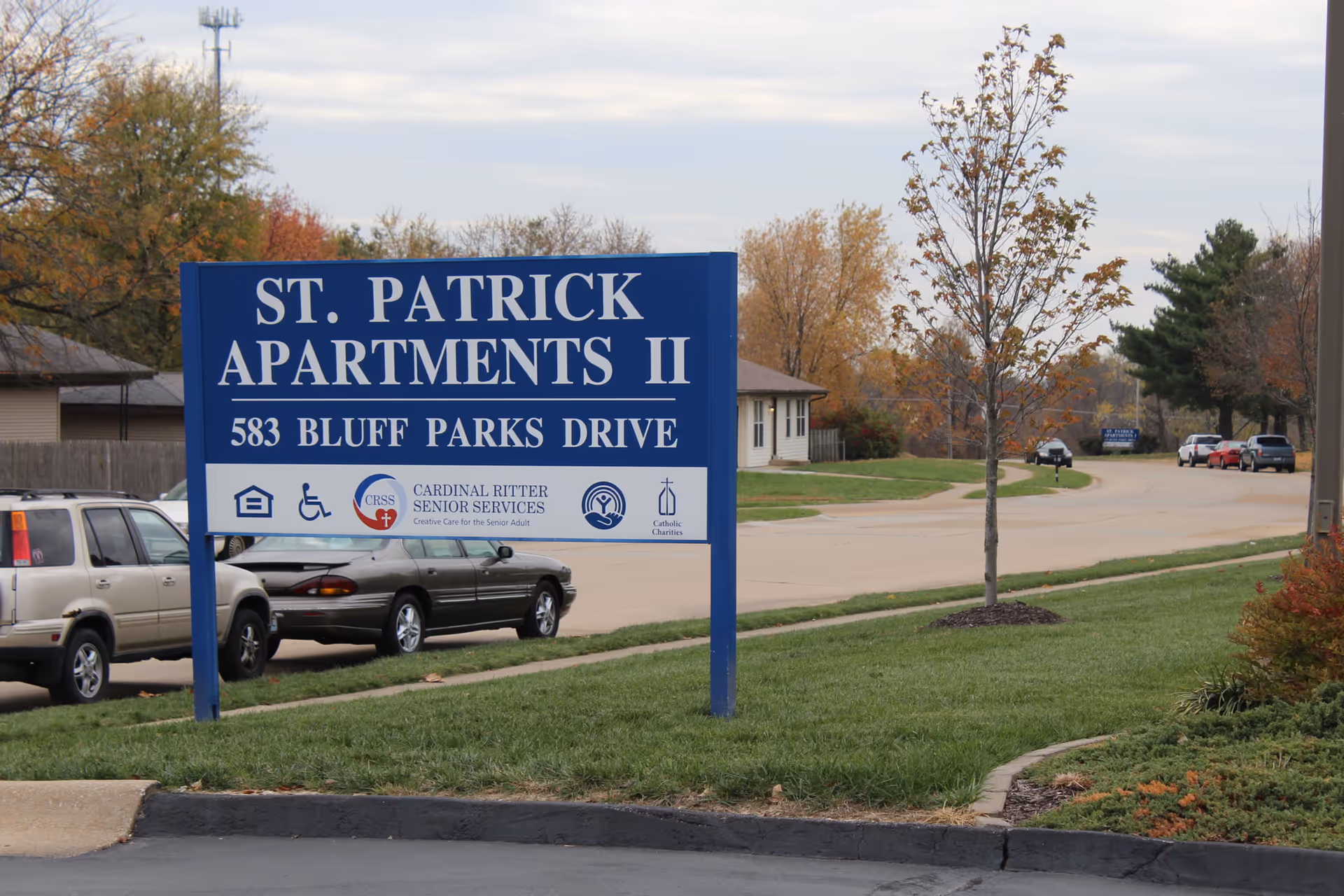 Blue sign reading St. Patrick Apartments II stands in a parking lot with trees.