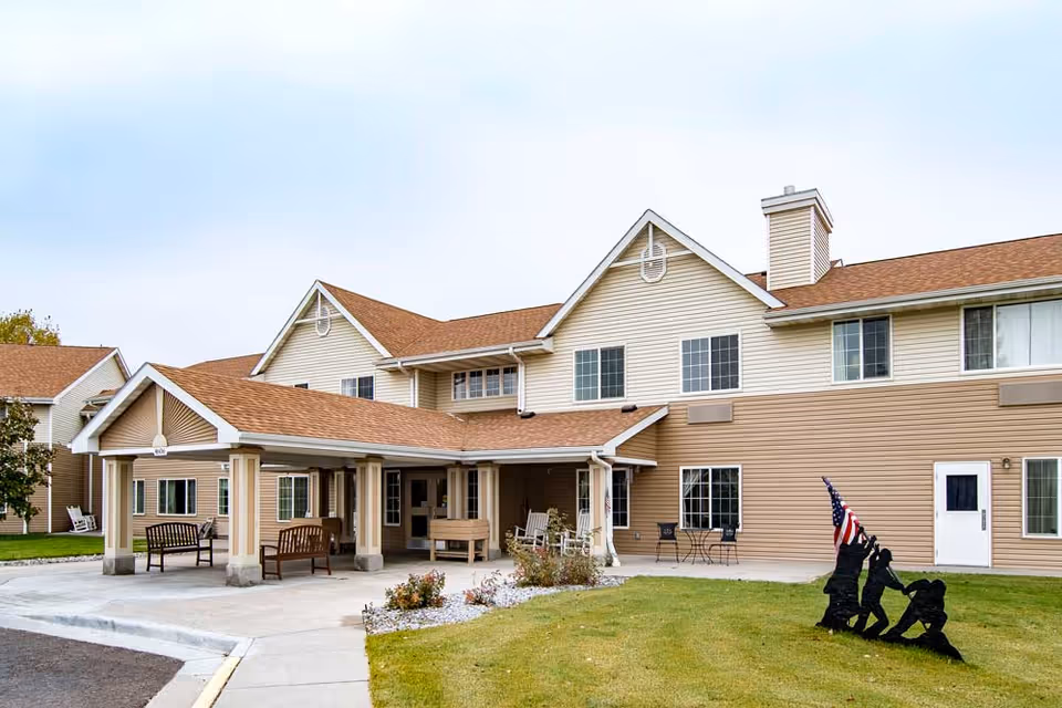 Two-story beige senior living building with a covered entrance, outdoor benches, and a lawn sculpture of soldiers raising an American flag.
