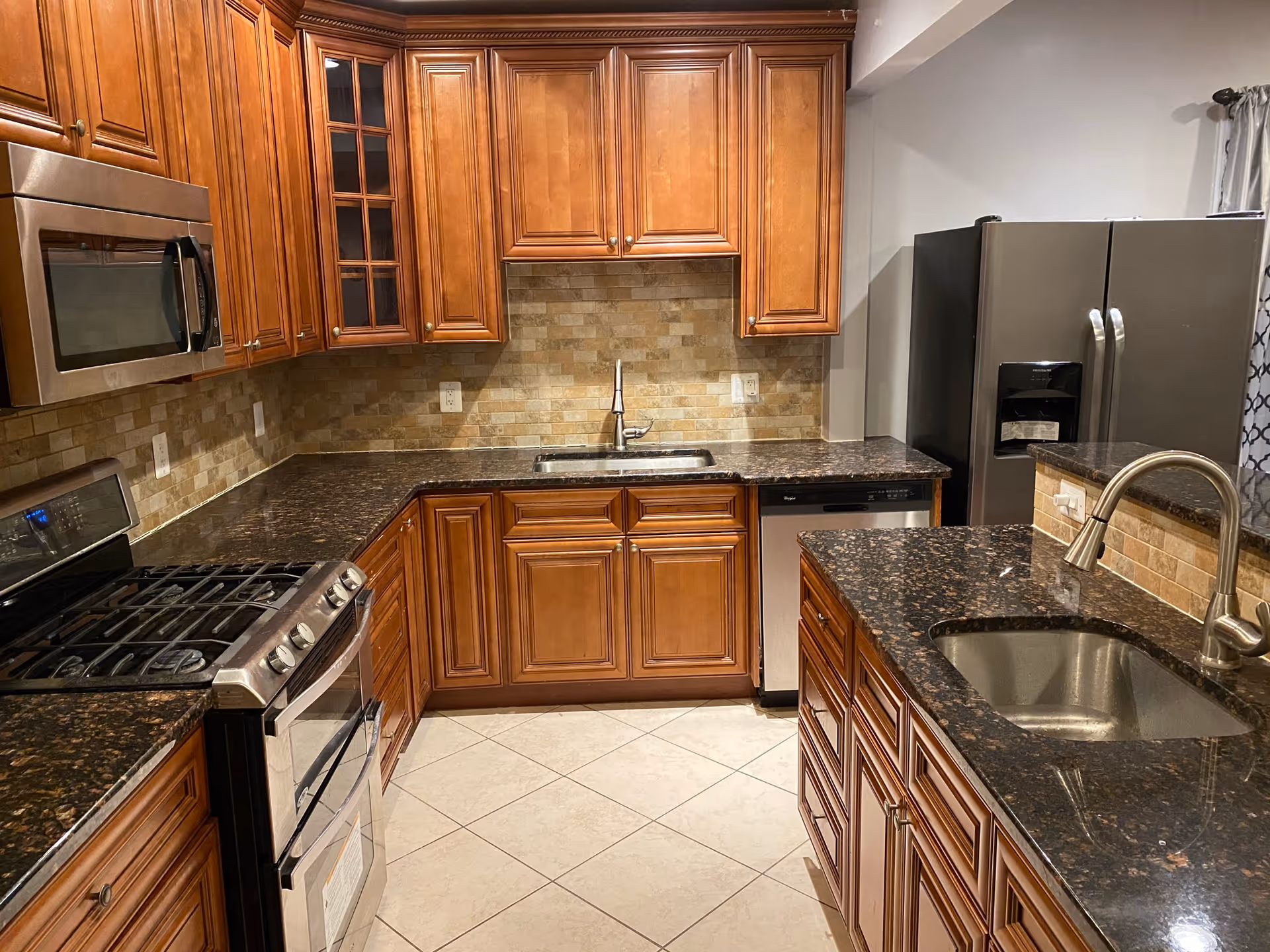 A modern kitchen with wooden cabinets, dark granite countertops, a stainless steel gas stove with oven, a microwave, a dishwasher, a double-door refrigerator, and two sinks with faucets. The backsplash is made of beige tiles, and the floor is tiled with light-colored tiles.