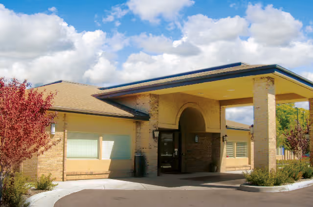 Exterior view of a single-story brick building with a covered entrance supported by two columns. There are small trees and shrubs planted around the building, and the sky is partly cloudy.