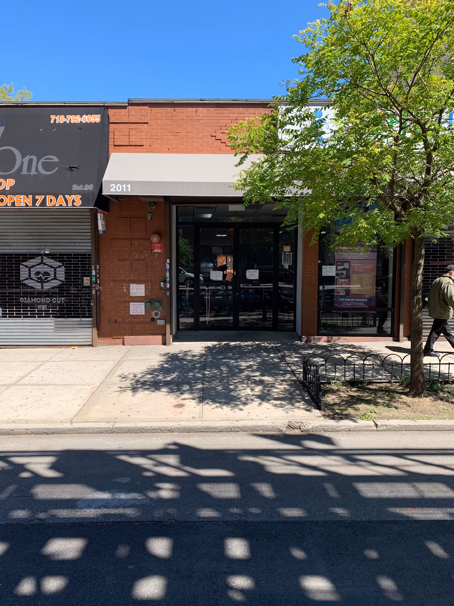 Brick storefront with glass doors, awning, and a tree on a sunny city sidewalk.