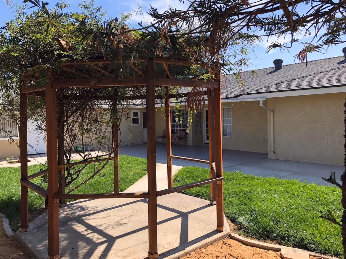 Outdoor area of a senior care facility featuring a wooden pergola with some climbing plants, a concrete pathway, green grass, and a beige building with windows and a covered patio in the background under a clear blue sky.