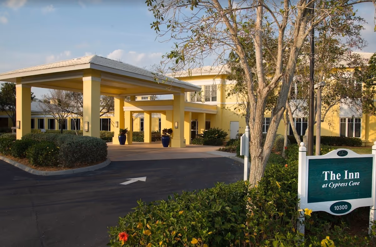 Entrance of a yellow two-story senior living building with a covered porte-cochère, landscaping, and a green sign reading "The Inn at Cypress Cove".