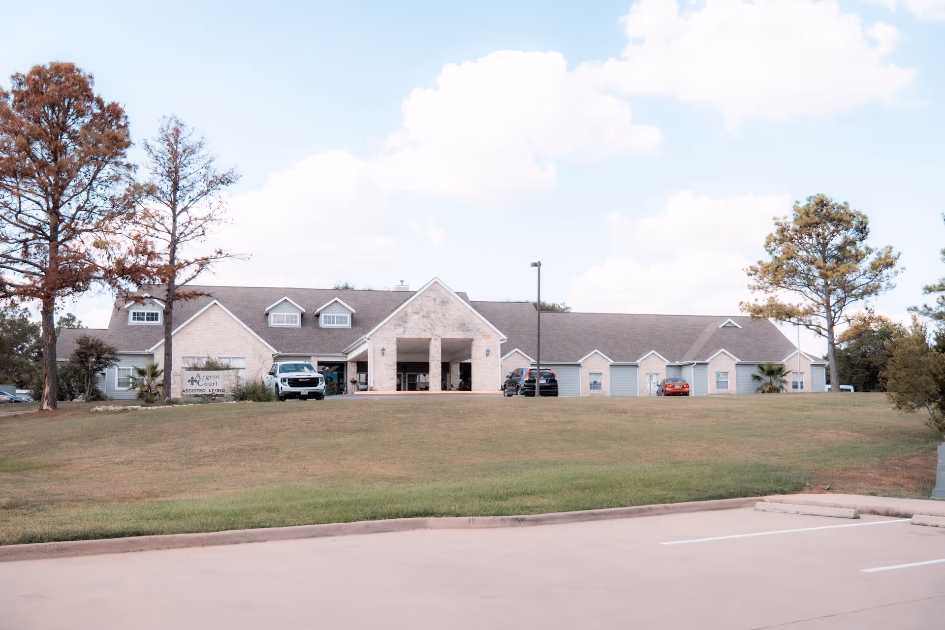 Front exterior view of Argent Court Assisted Living facility showing a single-story building with a stone facade, a covered entrance supported by columns, several parked cars, trees, and a grassy lawn under a partly cloudy sky.
