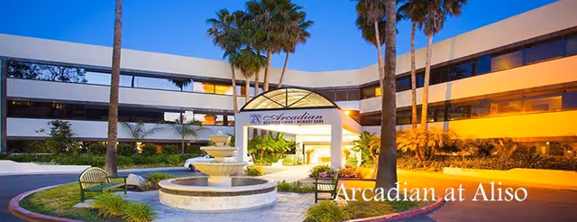Front exterior of the Arcadian at Aliso senior living building with a fountain, palm trees, and illuminated entrance canopy.