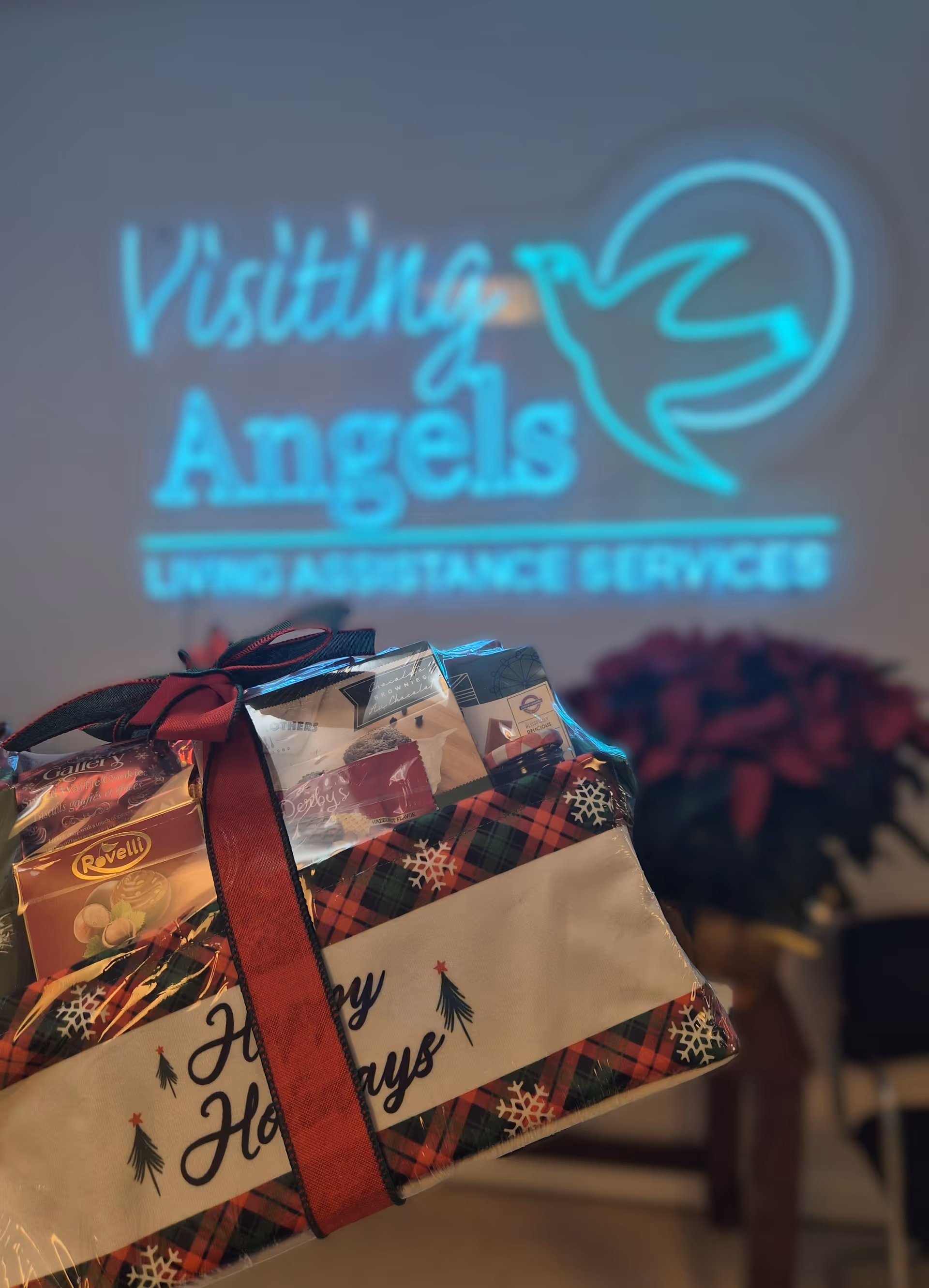 Gift box in foreground with neon Visiting Angels sign on a cozy indoor wall.