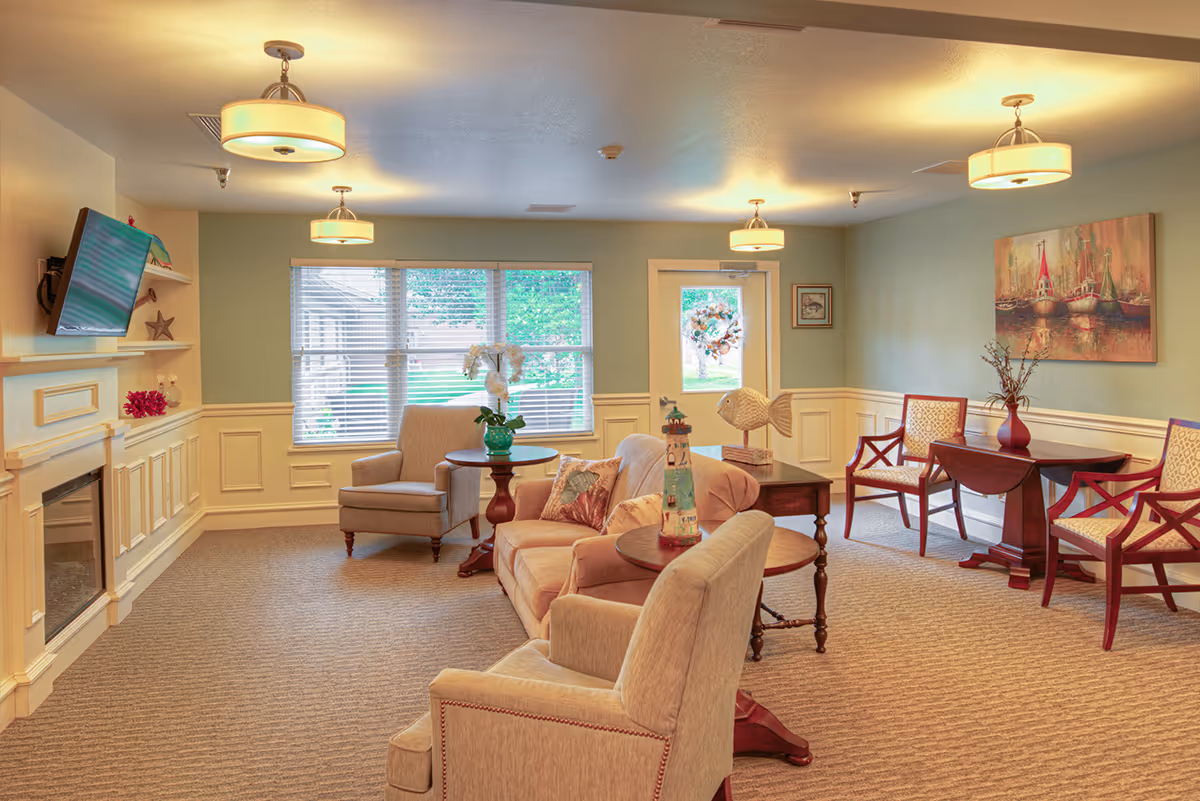 Well-lit communal living room with upholstered chairs, tables, a fireplace with a mounted TV, and decorative artwork.