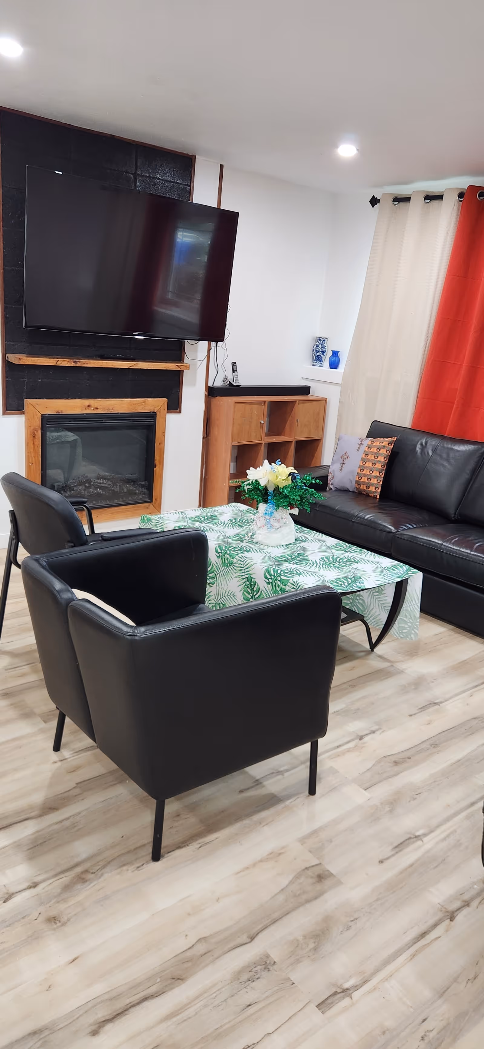 Modern living room with black leather seating, coffee table, fireplace, TV, and colorful curtains.