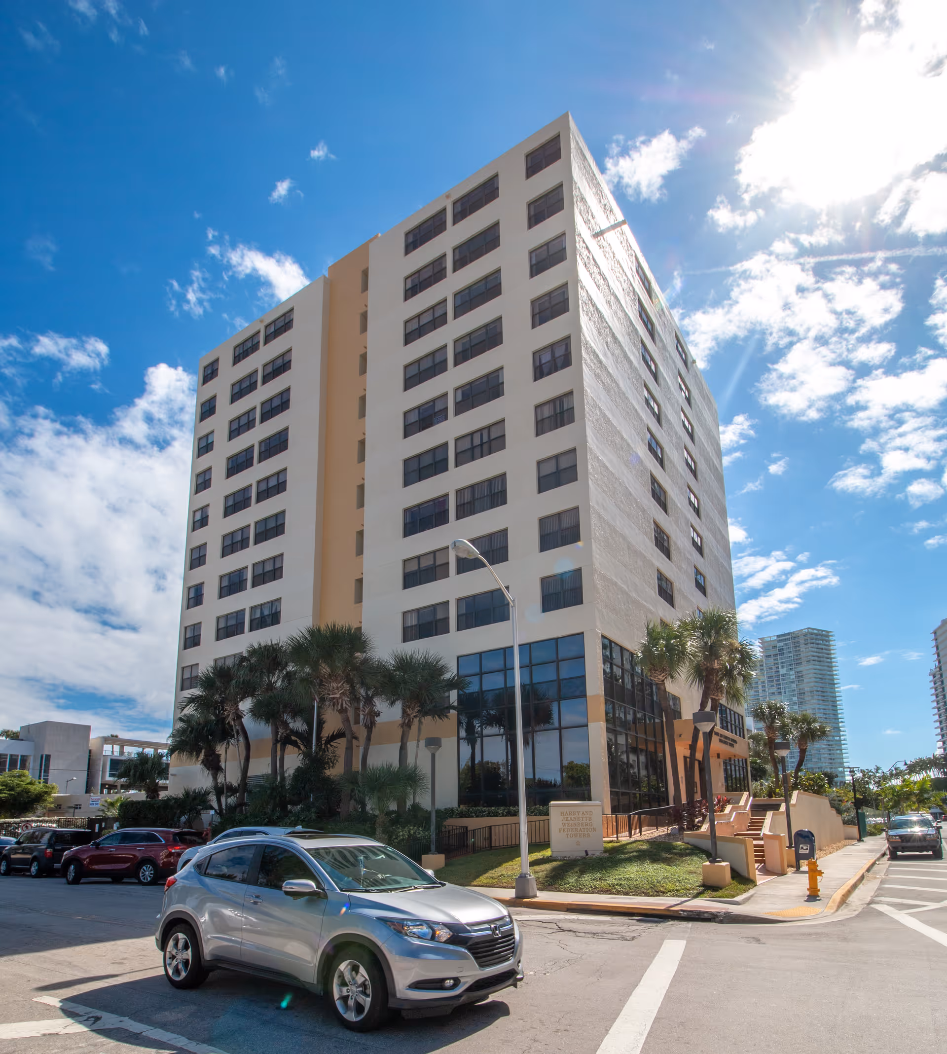 Tall, modern senior living tower with palm trees and street in front.