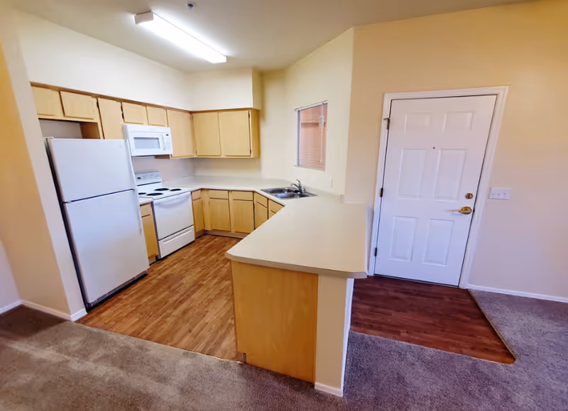Kitchen area with white appliances, light wood cabinets, and a doorway near a beige wall.