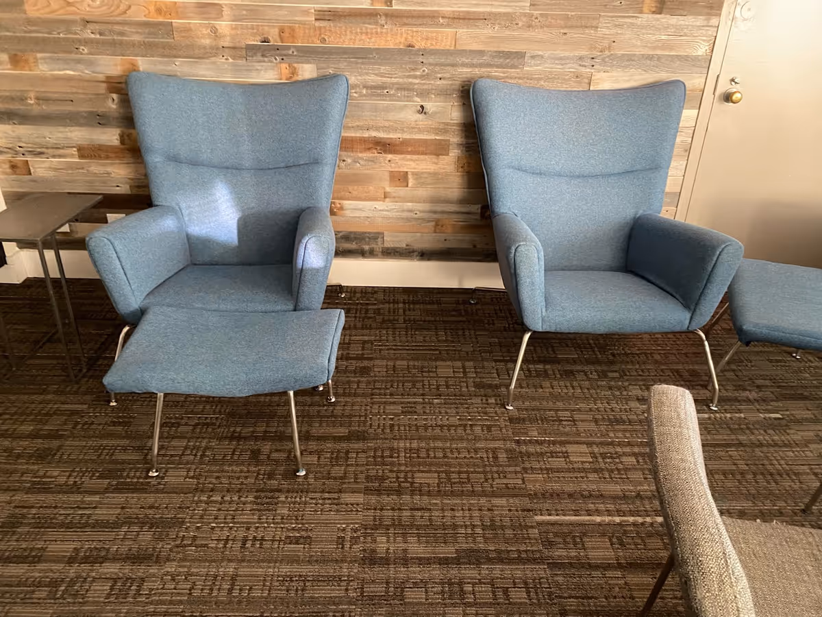 Two blue armchairs with an ottoman in a lounge area with a wood-paneled wall.