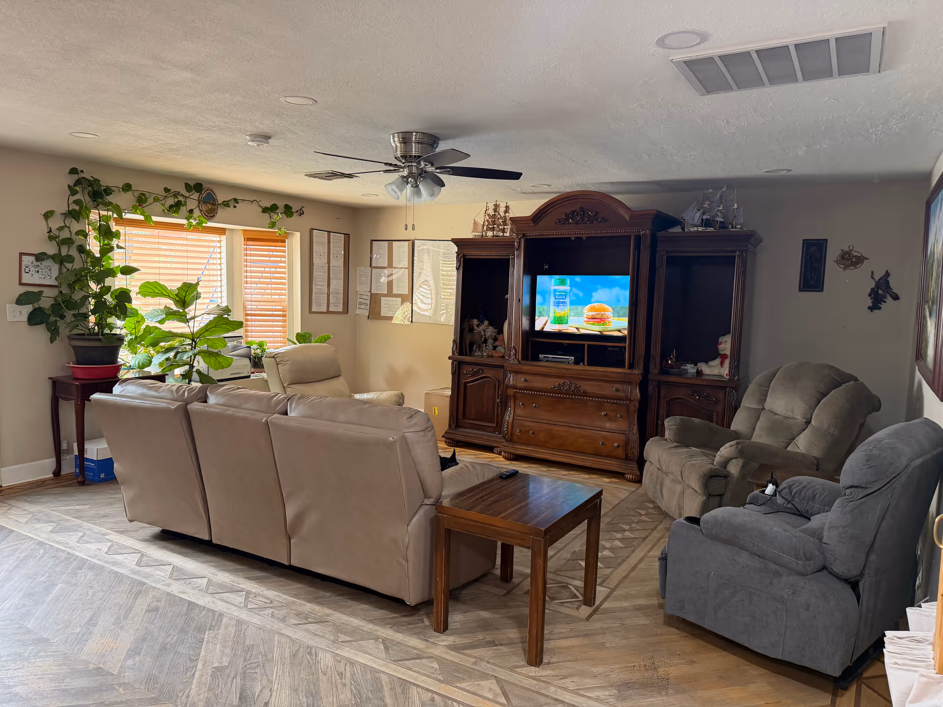 Cozy living room with beige couches, recliners, a wooden entertainment center, and potted plants.