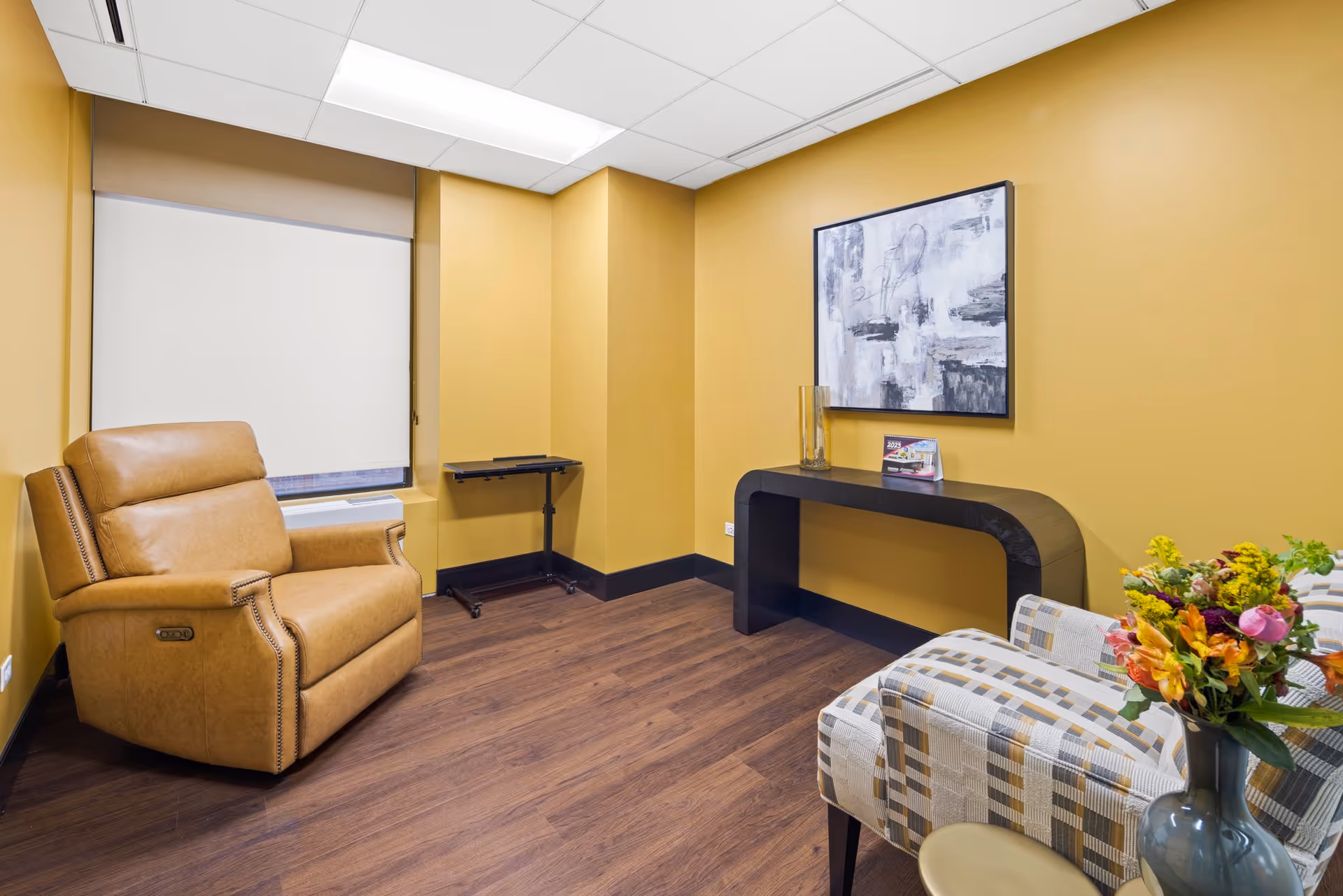 Waiting area in a facility with tan leather chair, desk, artwork, and flowers against yellow walls.