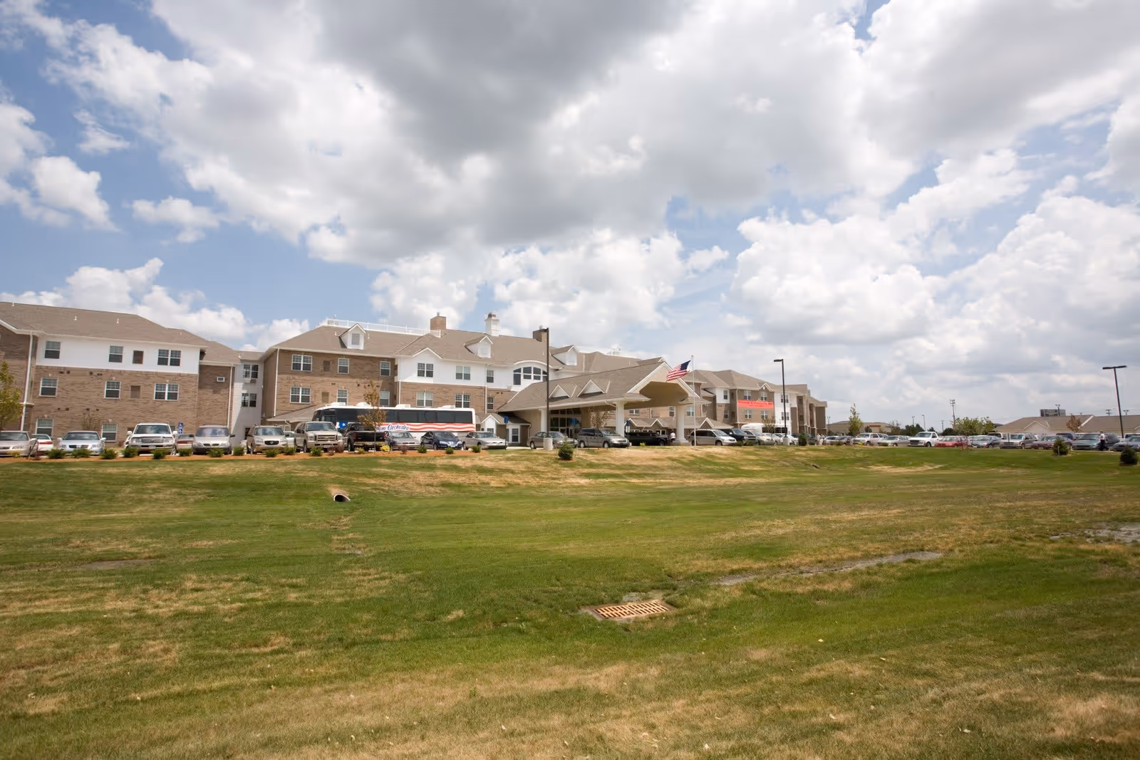 Exterior view of Marshall Square Retirement Community building with a large green lawn in the foreground, several parked cars, and a partly cloudy sky overhead.