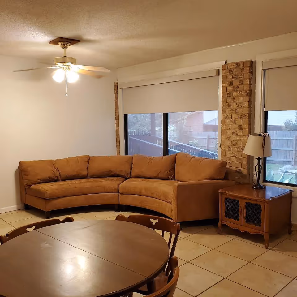 A cozy living room with a tan curved sectional sofa, round dining table in the foreground, large windows, and a side table with a lamp.