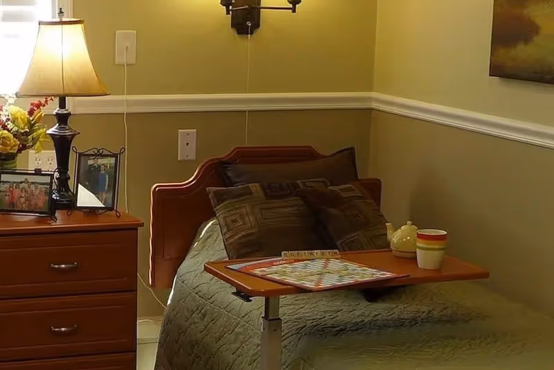 A cozy bedroom with a single bed covered in a green quilt and several decorative pillows. A wooden bedside table with a lamp, framed photos, and a flower arrangement is next to the bed. A small adjustable table on the bed holds a Scrabble game, a teapot, and a cup. The walls are painted beige with white trim, and a painting hangs on the wall.