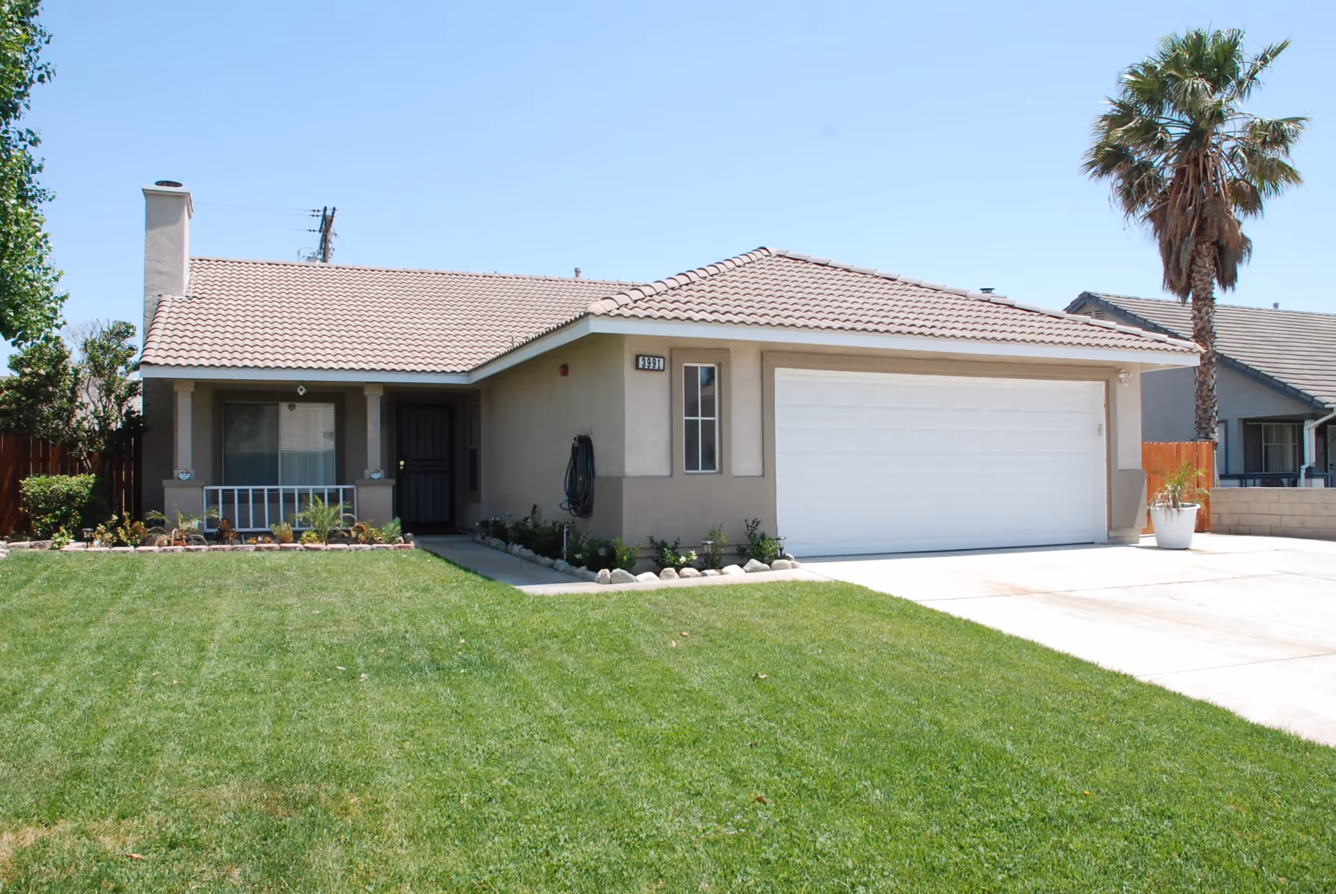 Single-story house with a tiled roof, a well-maintained green lawn, a driveway leading to a white garage door, a small front porch with a black door, and a palm tree on the right side under a clear blue sky.