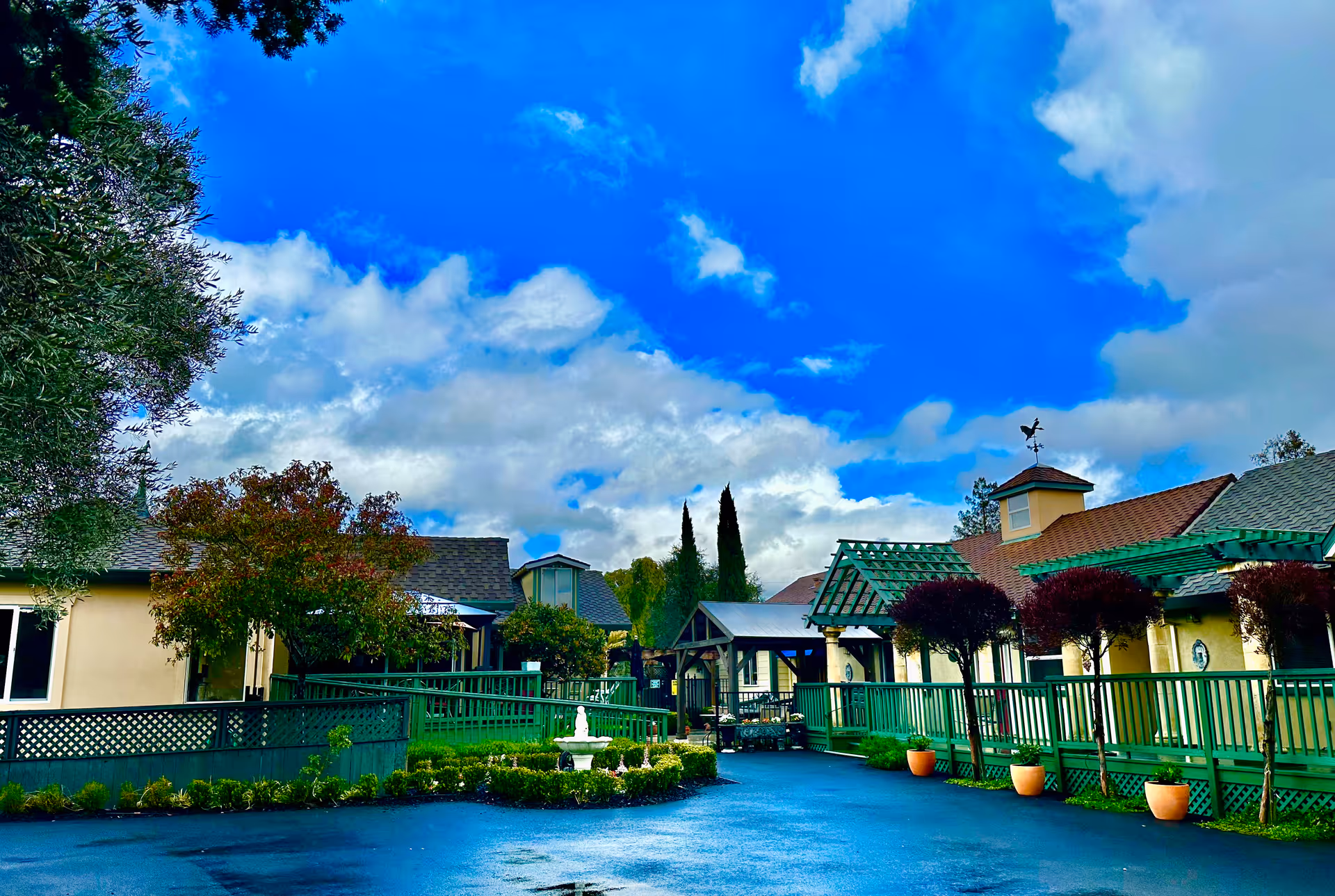 Outdoor view of Bella Vista Village II showing a courtyard with green railings, potted plants, trees, a small fountain, and buildings with pitched roofs under a partly cloudy blue sky.