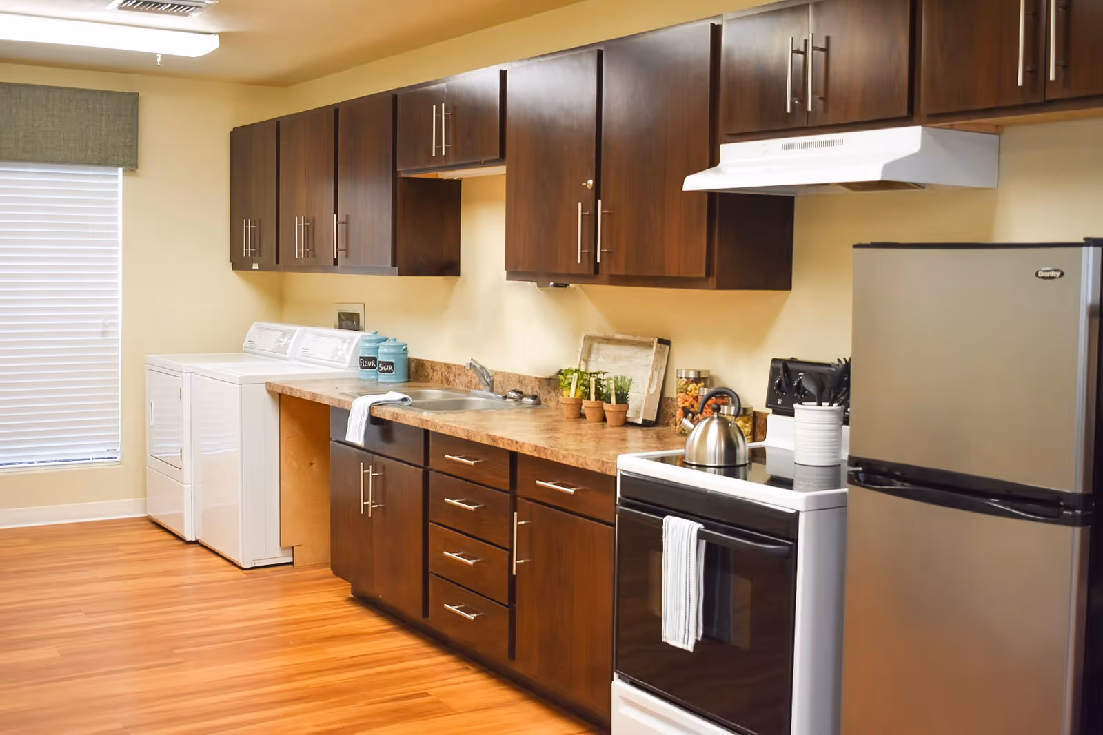A clean and modern kitchen area with dark wood cabinets, a countertop with a sink, a stainless steel kettle on a stove, a small refrigerator, and a washer and dryer set near a window with blinds.