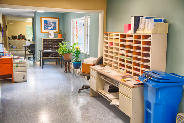 Indoor facility office area with filing shelves, desks, plants, and a blue recycling bin.