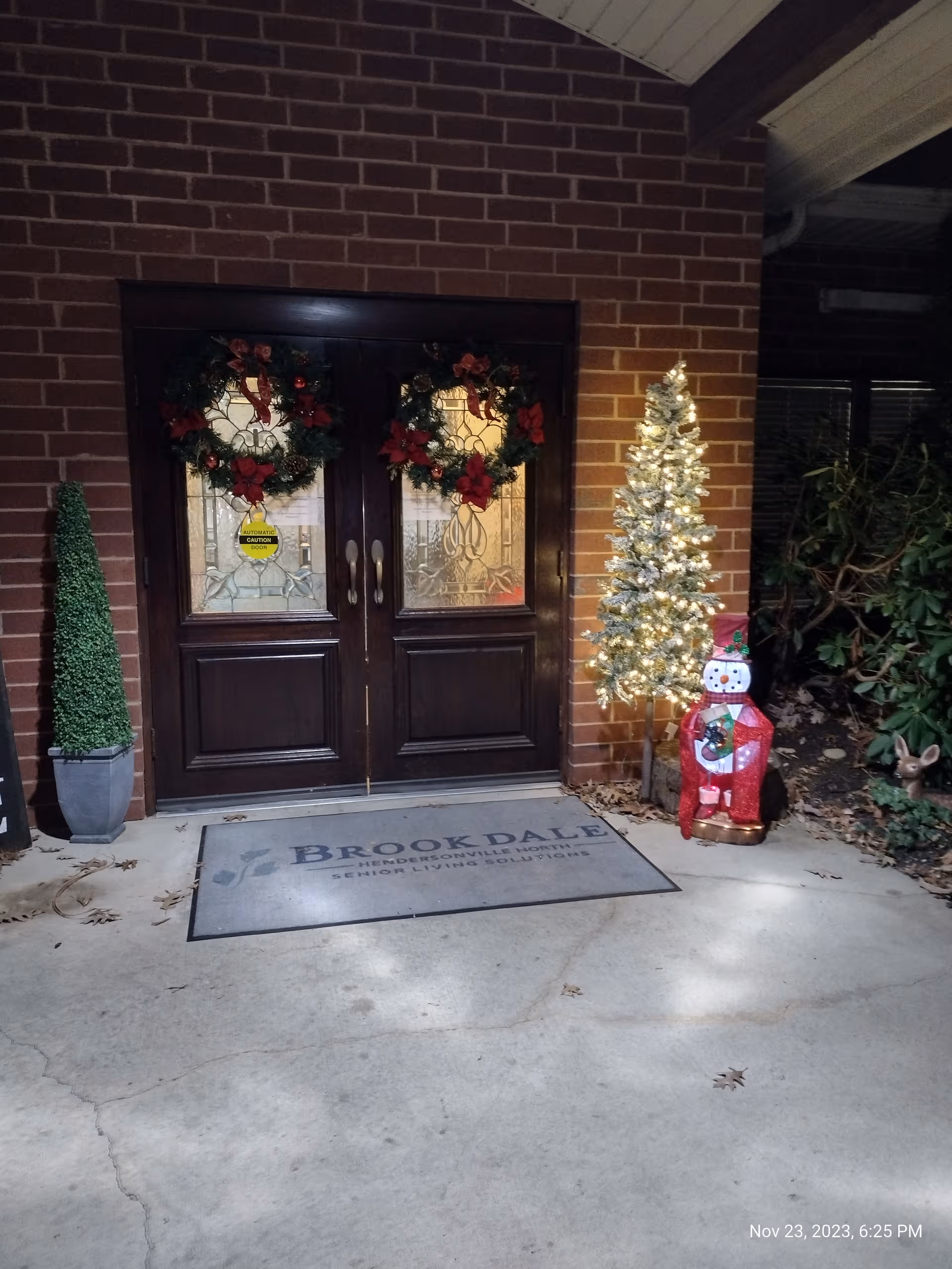 Double wooden doors with holiday wreaths, a lit Christmas tree, and a Brookdale mat outside.