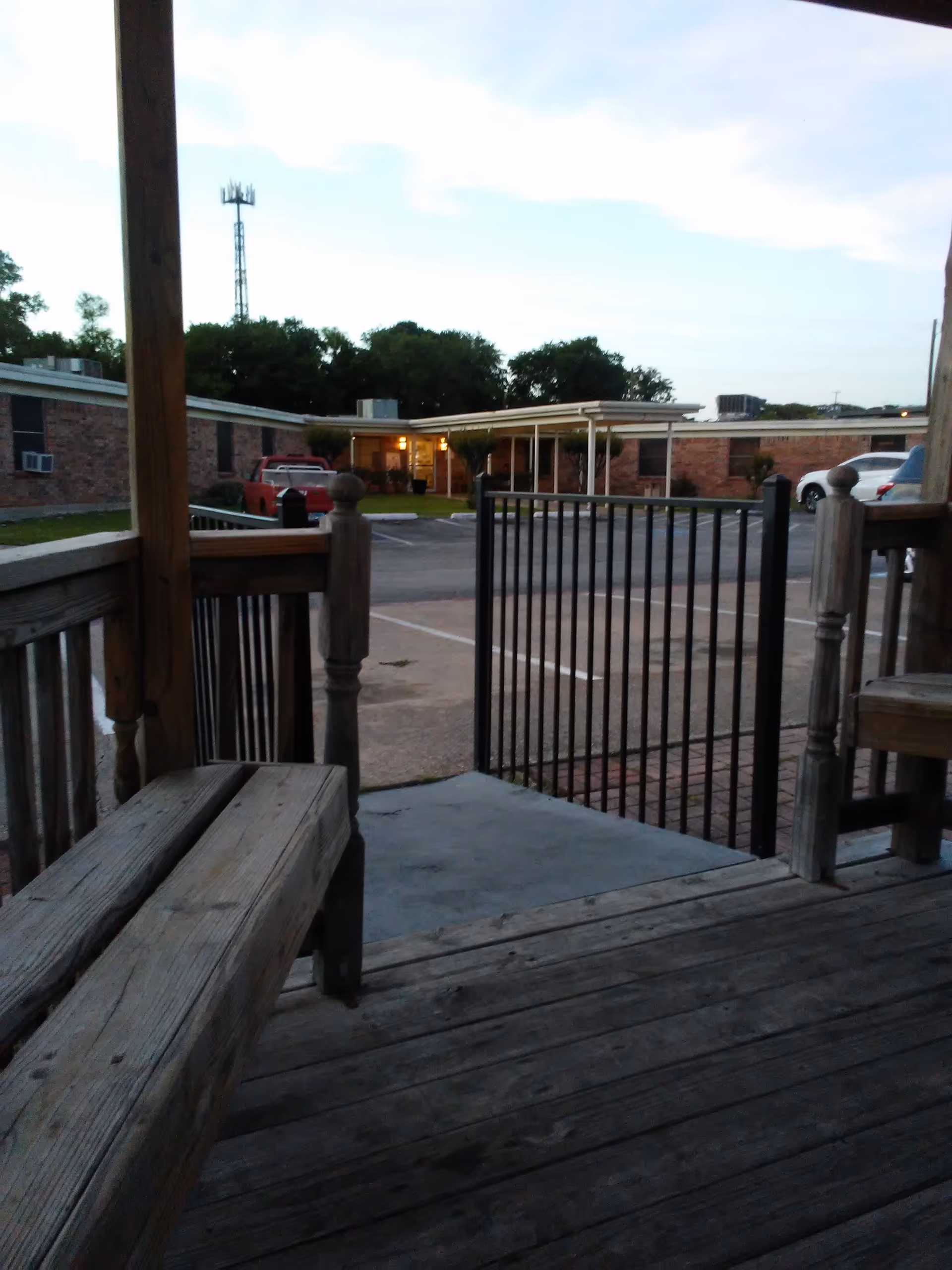 Wooden porch with bench and railing overlooking a parking lot and brick buildings.