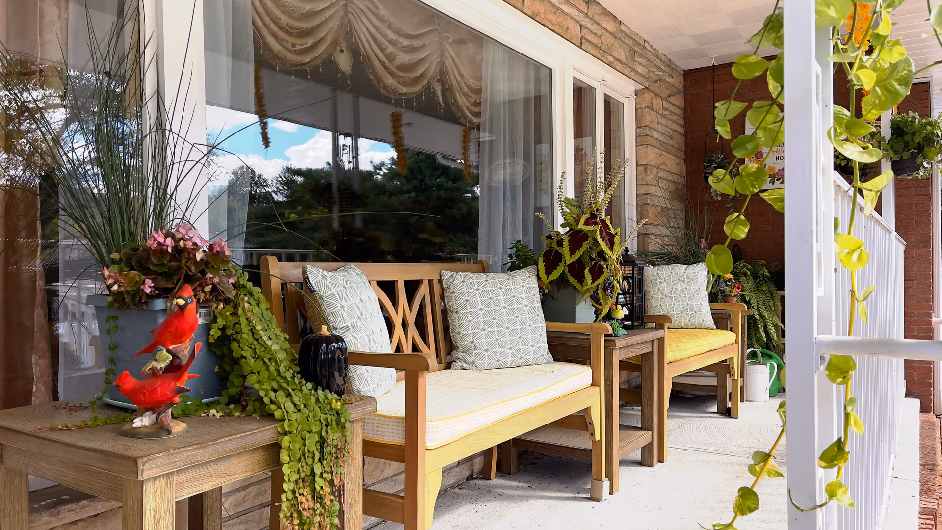 A cozy outdoor porch area with wooden benches featuring yellow cushions and patterned pillows. The porch is decorated with various green plants and flowers, including hanging vines and potted plants on small wooden tables. The background shows a large window with curtains and a brick wall.