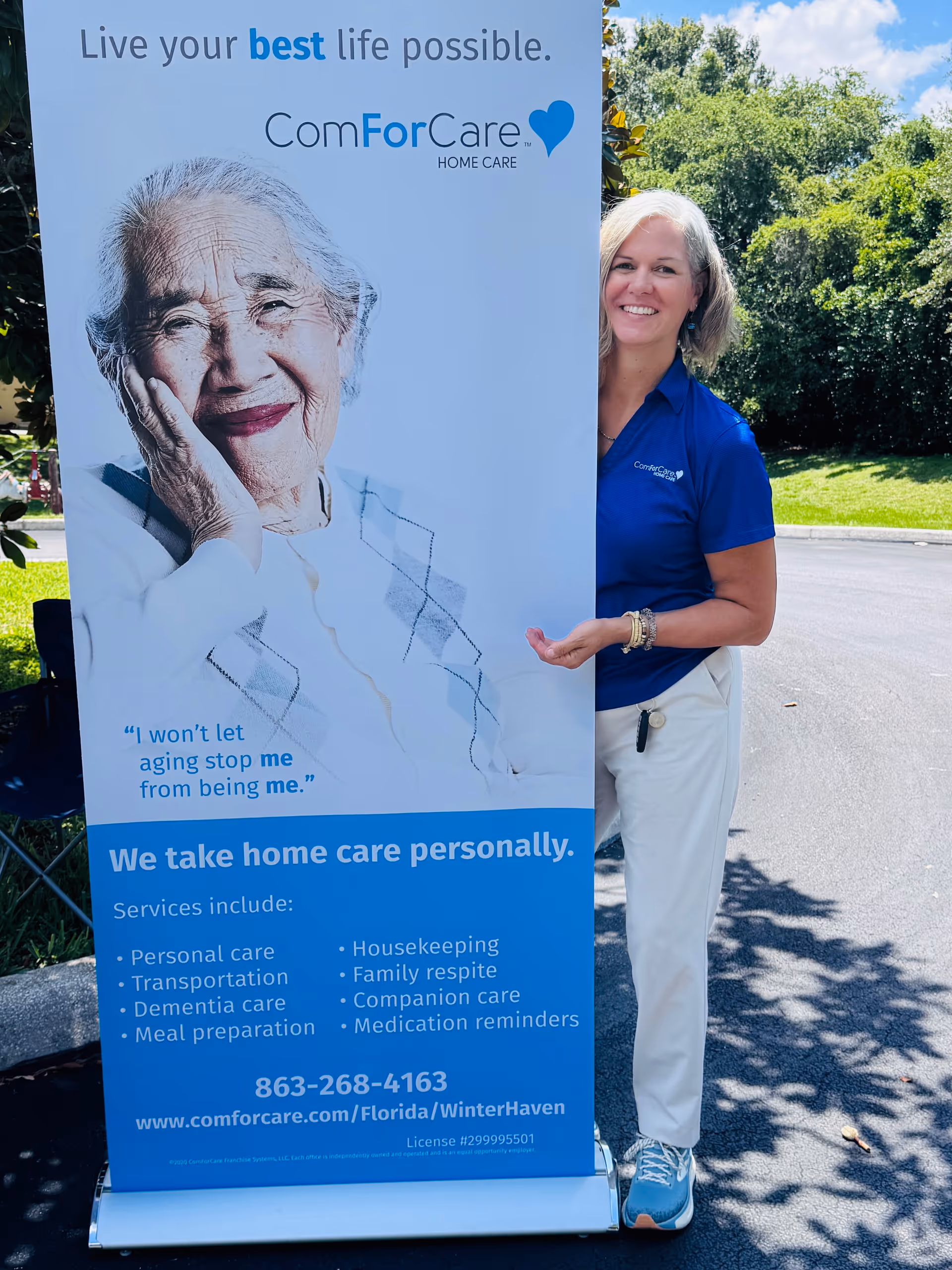 Outdoor scene with a ComForCare banner and a woman in blue polo standing beside it.