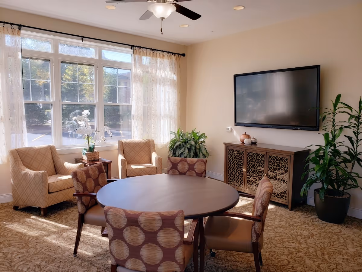 Cozy lounge area with a round table, chairs, plants, and a wall-mounted TV.
