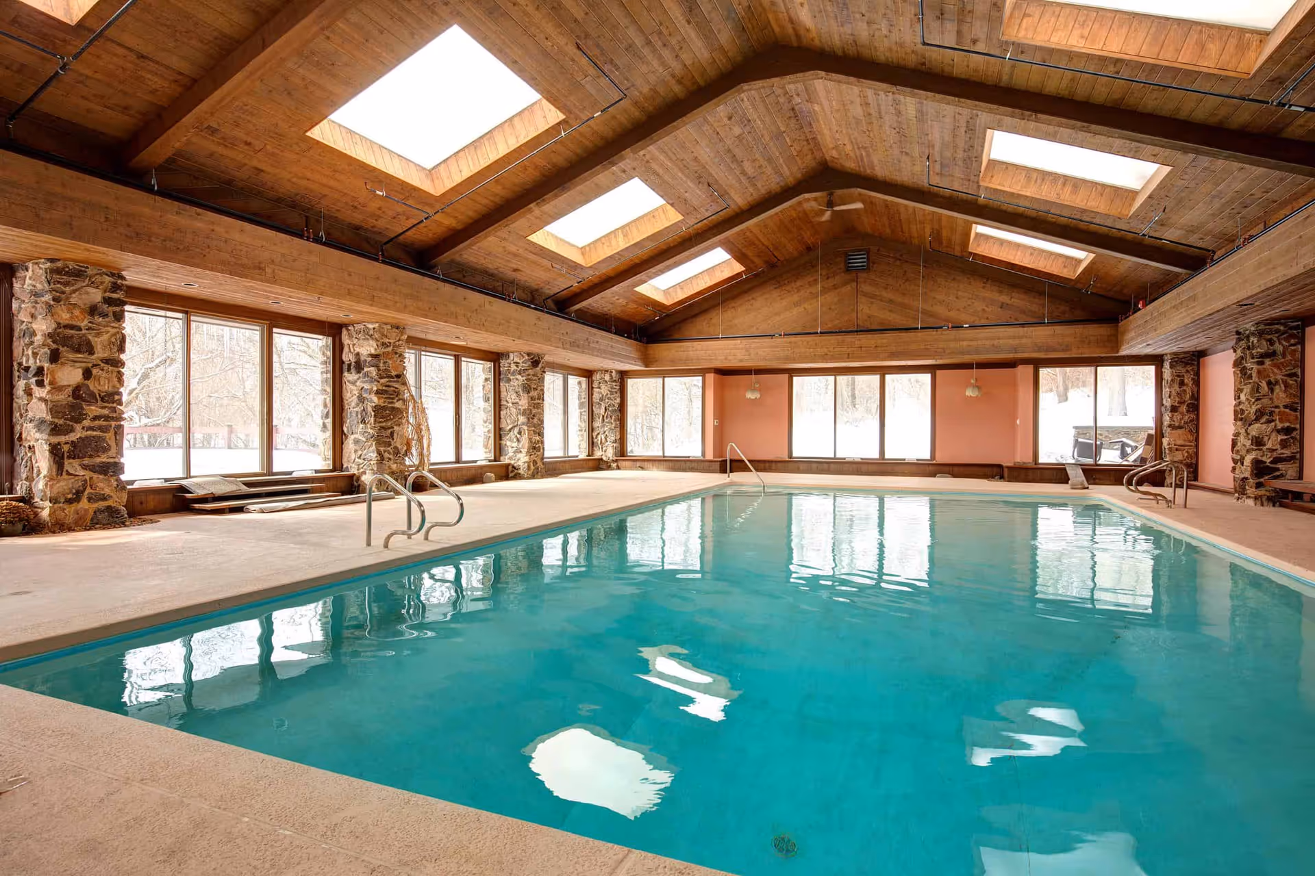 Indoor swimming pool in a wood-paneled room with skylights, large windows, and stone columns.