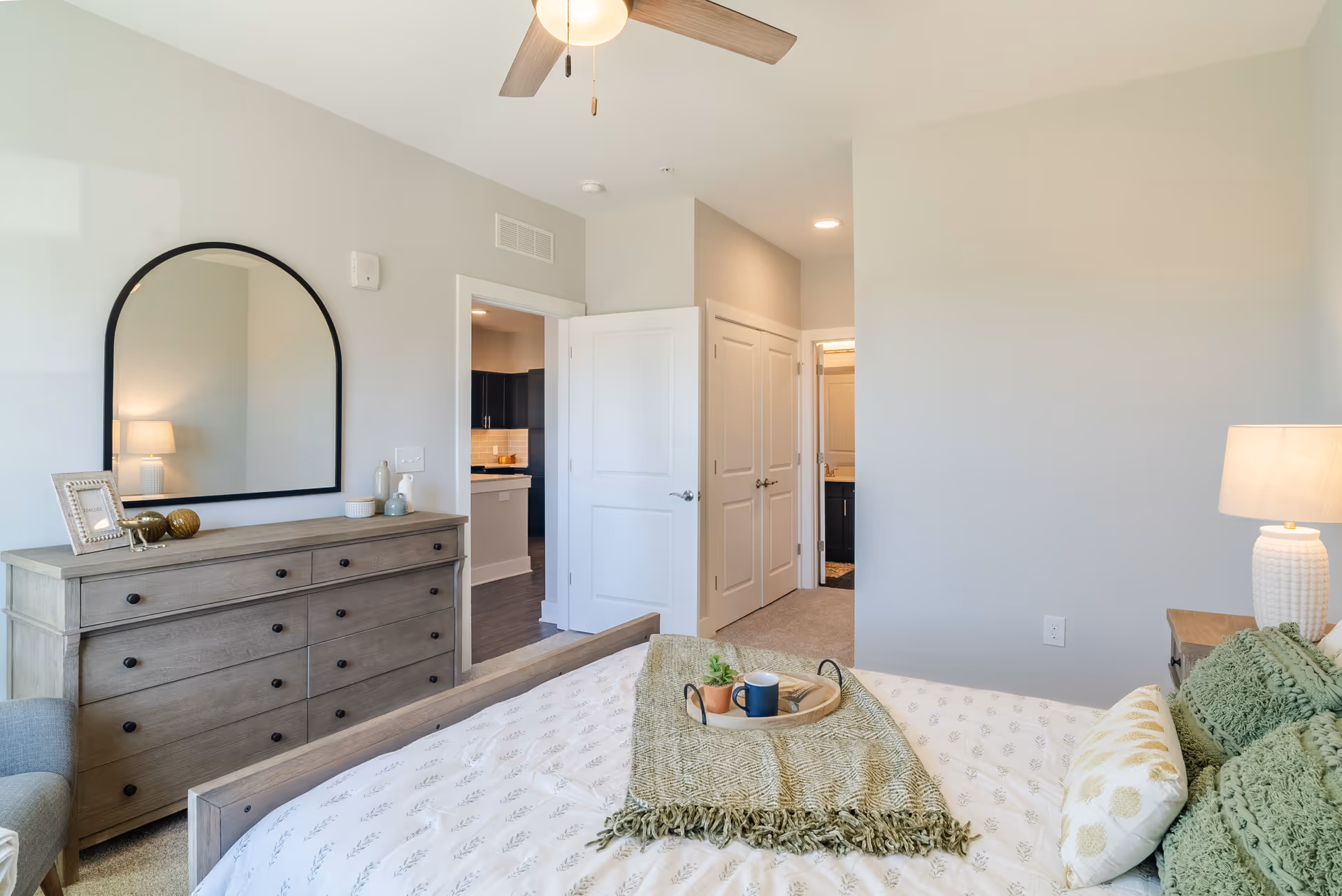 A cozy bedroom with a dresser, arched mirror, bed, lamp, and doorway to the kitchen.