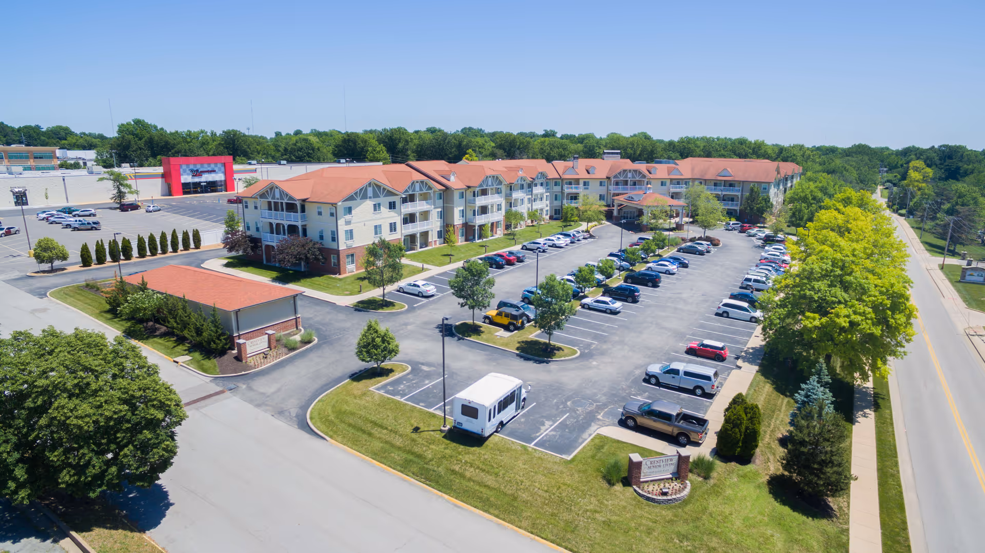 Aerial view of Crestview Senior Living facility showing a large three-story building with red roofs, multiple balconies, and a spacious parking lot filled with cars. The surrounding area includes trees, a road, and a small sign near the entrance.