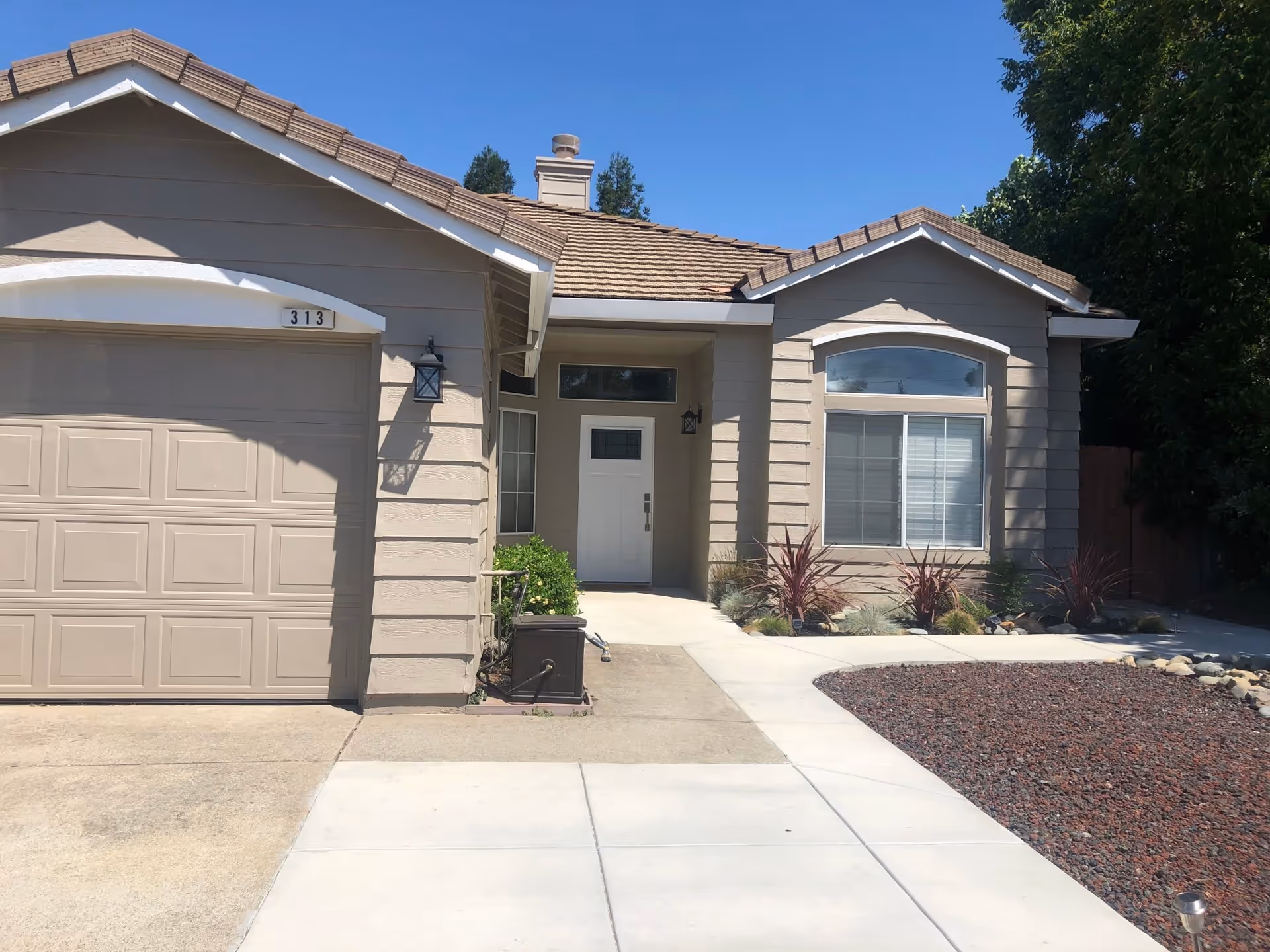Front exterior of a beige single-story house with a garage, driveway, and small landscaped yard.