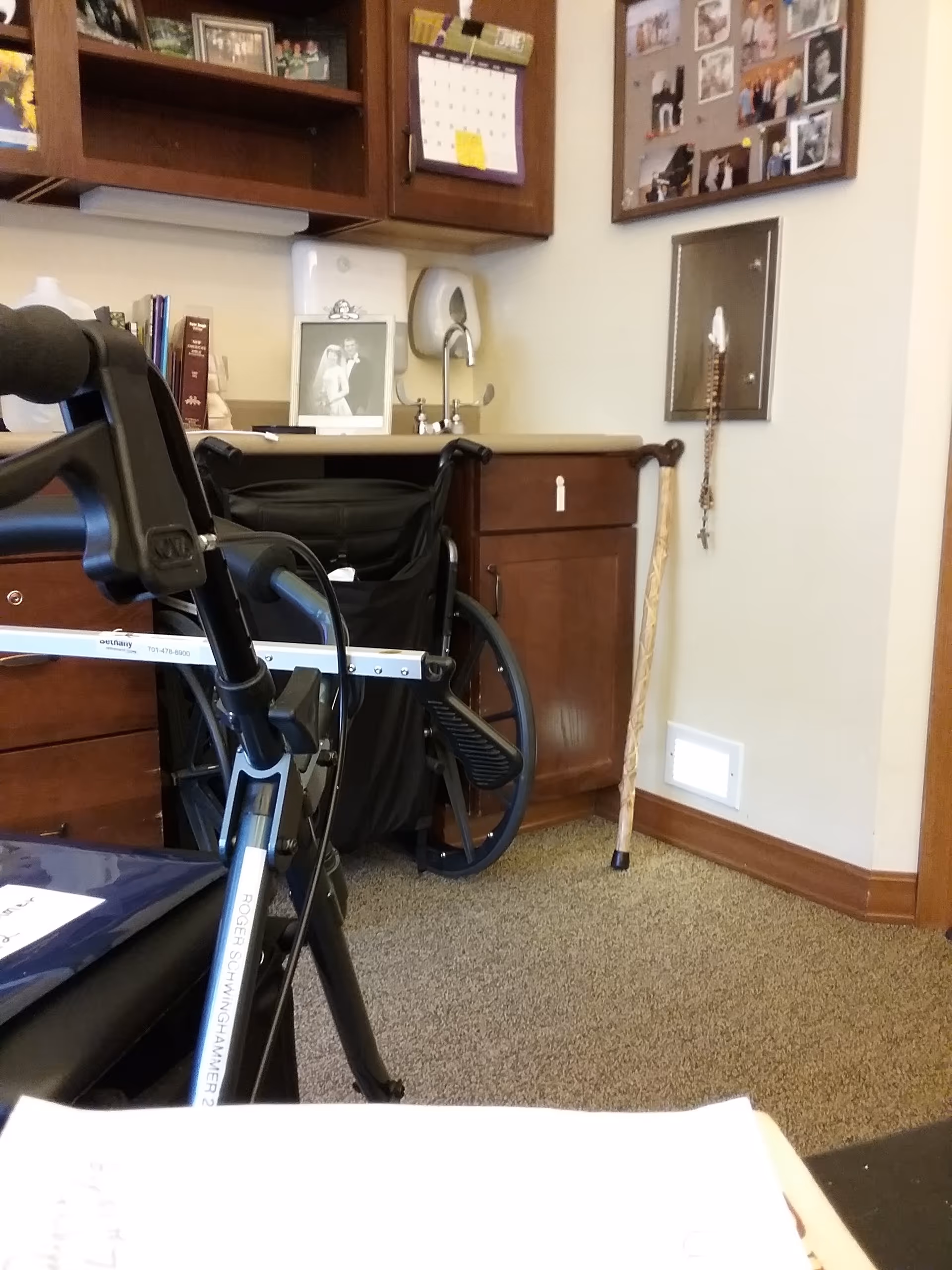 Wheelchair beside a desk with cabinets, photos, and a cane in an office-like room.