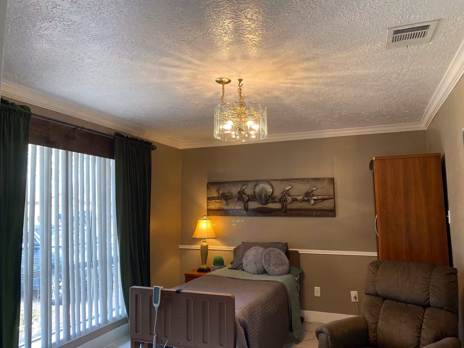 A furnished bedroom in a senior living facility with a bed, lamp, armoire, and curtains.