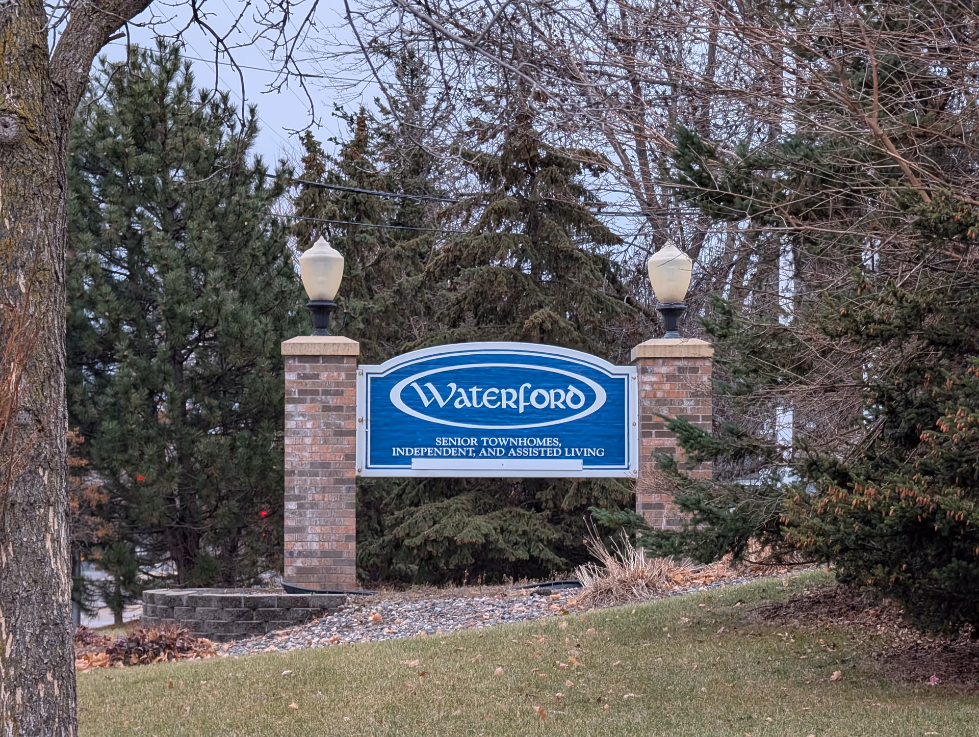 Blue 'Waterford' entrance sign for a senior living community mounted between brick pillars with lamp posts, surrounded by trees and grass.