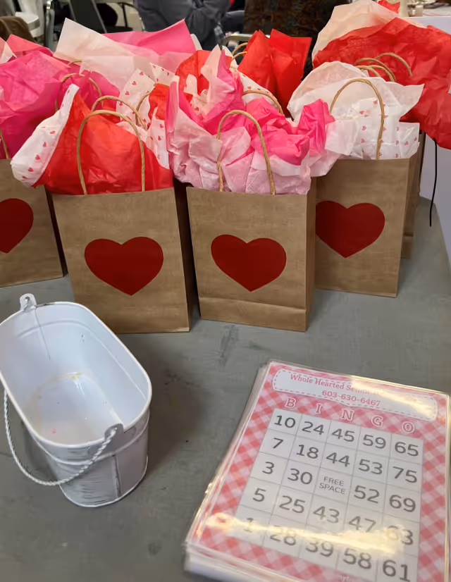 Brown gift bags with red hearts and pink tissue beside a bingo card on a table.