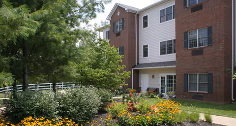 Exterior view of a multi-story senior living facility building with brick and white siding, surrounded by a landscaped garden with colorful flowers, bushes, and trees under a clear sky.