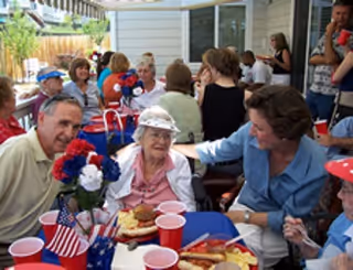 A group of elderly people and caregivers gathered outdoors around tables decorated with patriotic-themed centerpieces and red, white, and blue cups. They are enjoying a meal together in a social setting with a festive atmosphere.