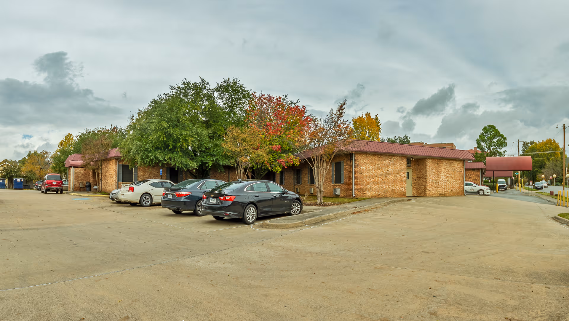 Single-story brick nursing facility building with a covered entrance, parked cars, and trees with autumn foliage in a large front parking lot.