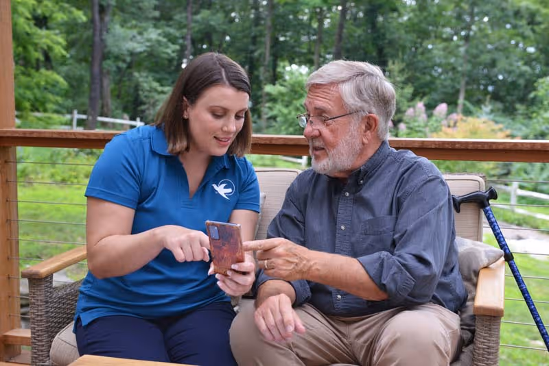 Caregiver in a blue polo assists an elderly man on a wooden outdoor porch.