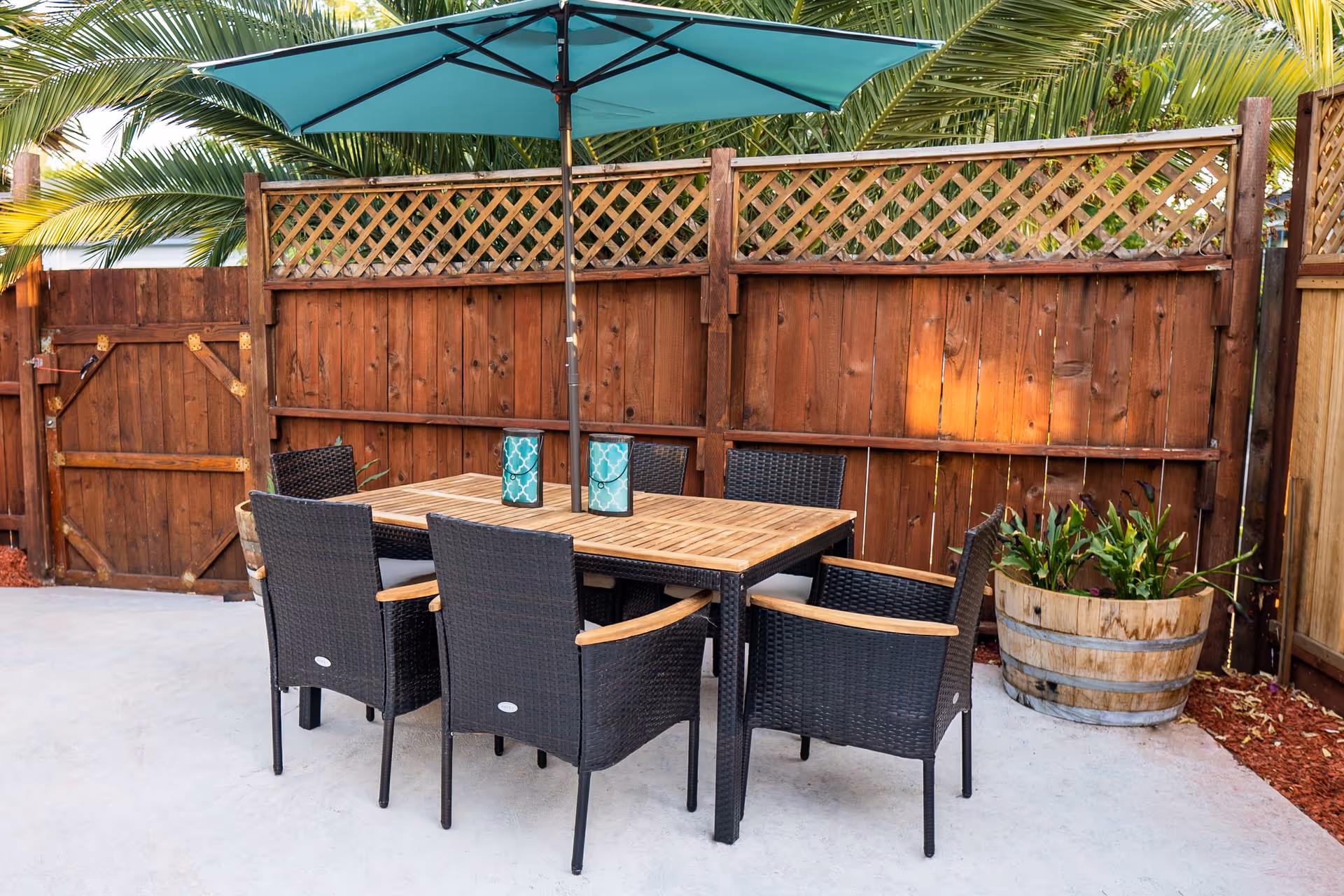 Outdoor patio area with a wooden table and six black wicker chairs with armrests. A large green umbrella is positioned in the center of the table. The area is enclosed by a wooden fence with lattice panels on top, and there is a large wooden planter with green plants on the right side.