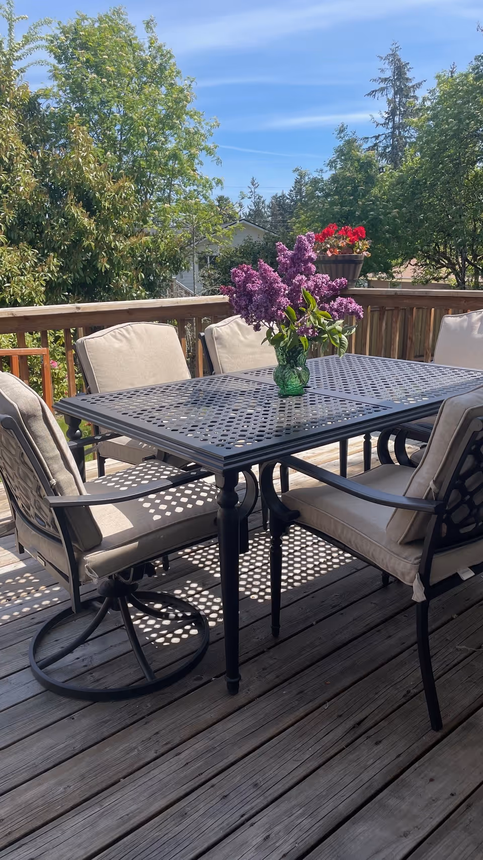 Outdoor wooden deck with a black metal table and four cushioned chairs. A green vase with purple flowers is placed on the table. Surrounding the deck are green trees and a clear blue sky.