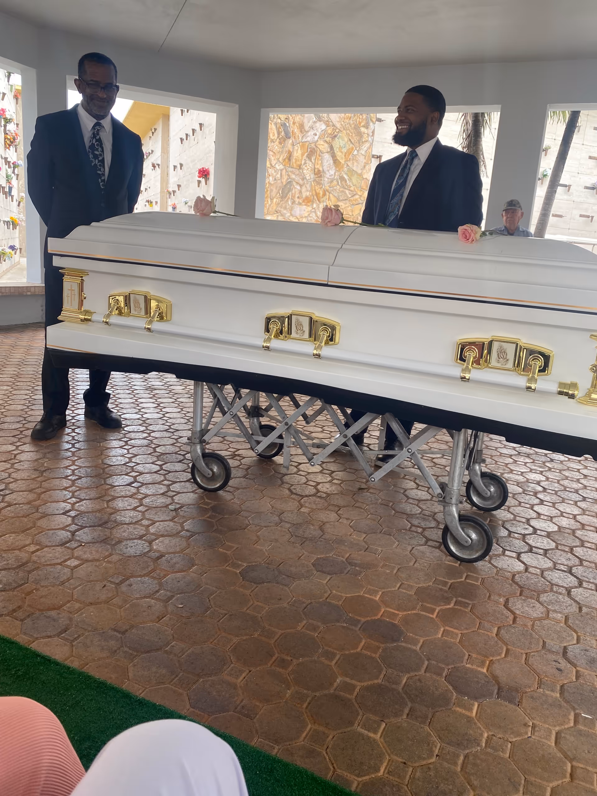 Two men in suits stand beside a white coffin on a wheeled bier outdoors.