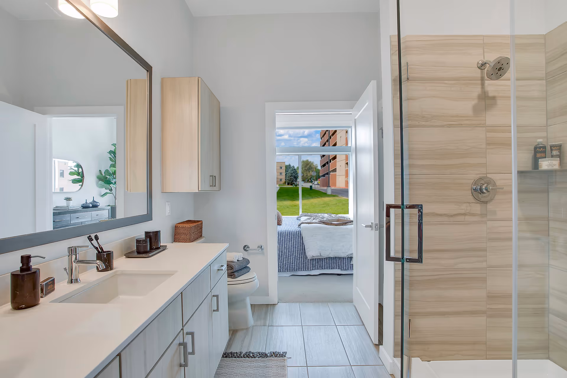 Modern bathroom with a large mirror above a white countertop and sink, light wood cabinets, a toilet with folded towels on top, and a glass-enclosed shower with beige tiles. The bathroom door is open, showing a bedroom with a bed and a large window overlooking a green lawn and buildings outside.