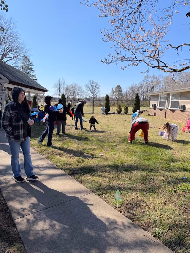Group of residents and children outdoors near a building with blooming trees on a sunny day.