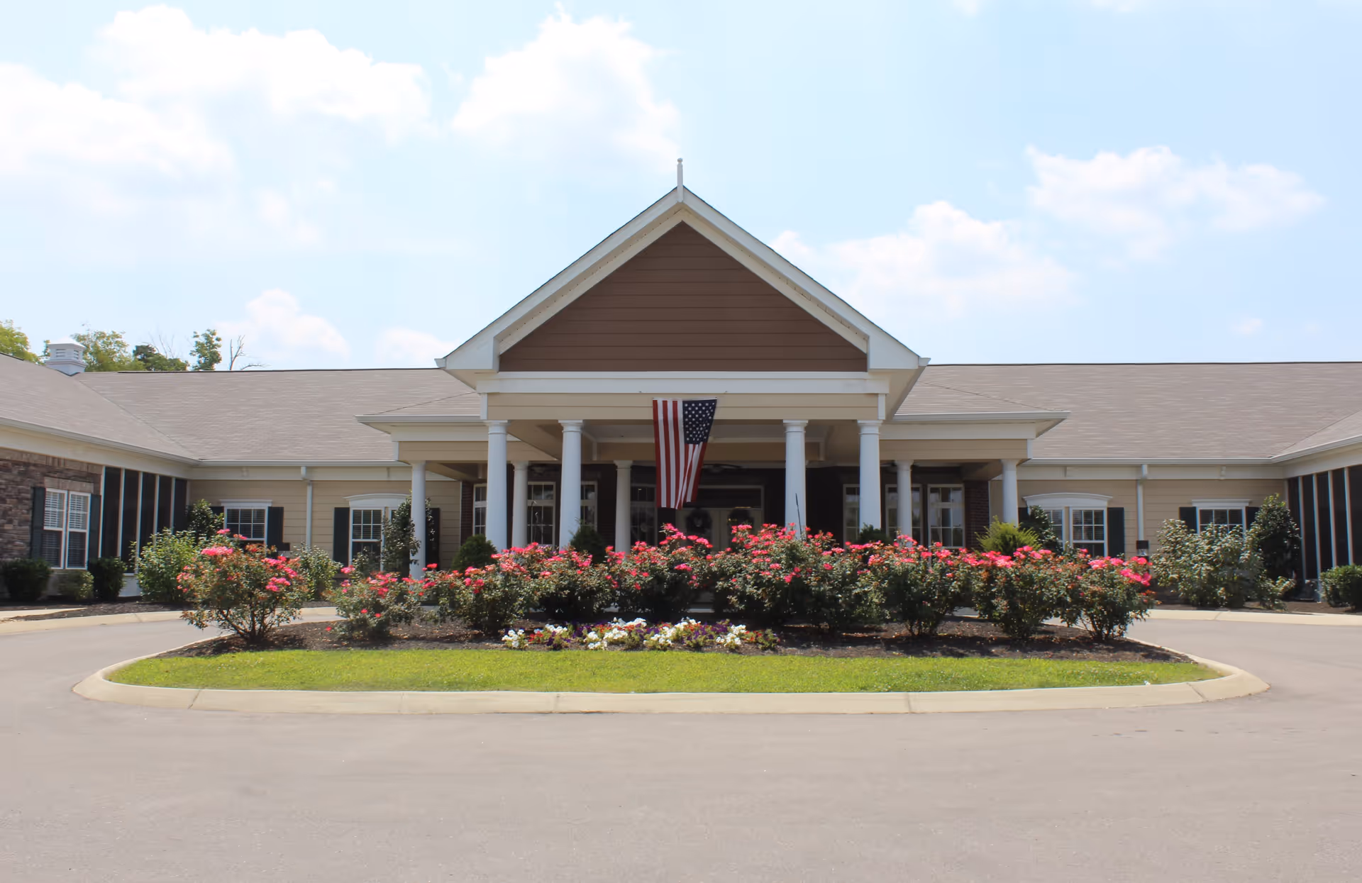Front exterior view of Carrick Glen Senior Living facility with a circular driveway, landscaped flower beds with pink and white flowers, white columns supporting a peaked roof, and an American flag hanging above the entrance.