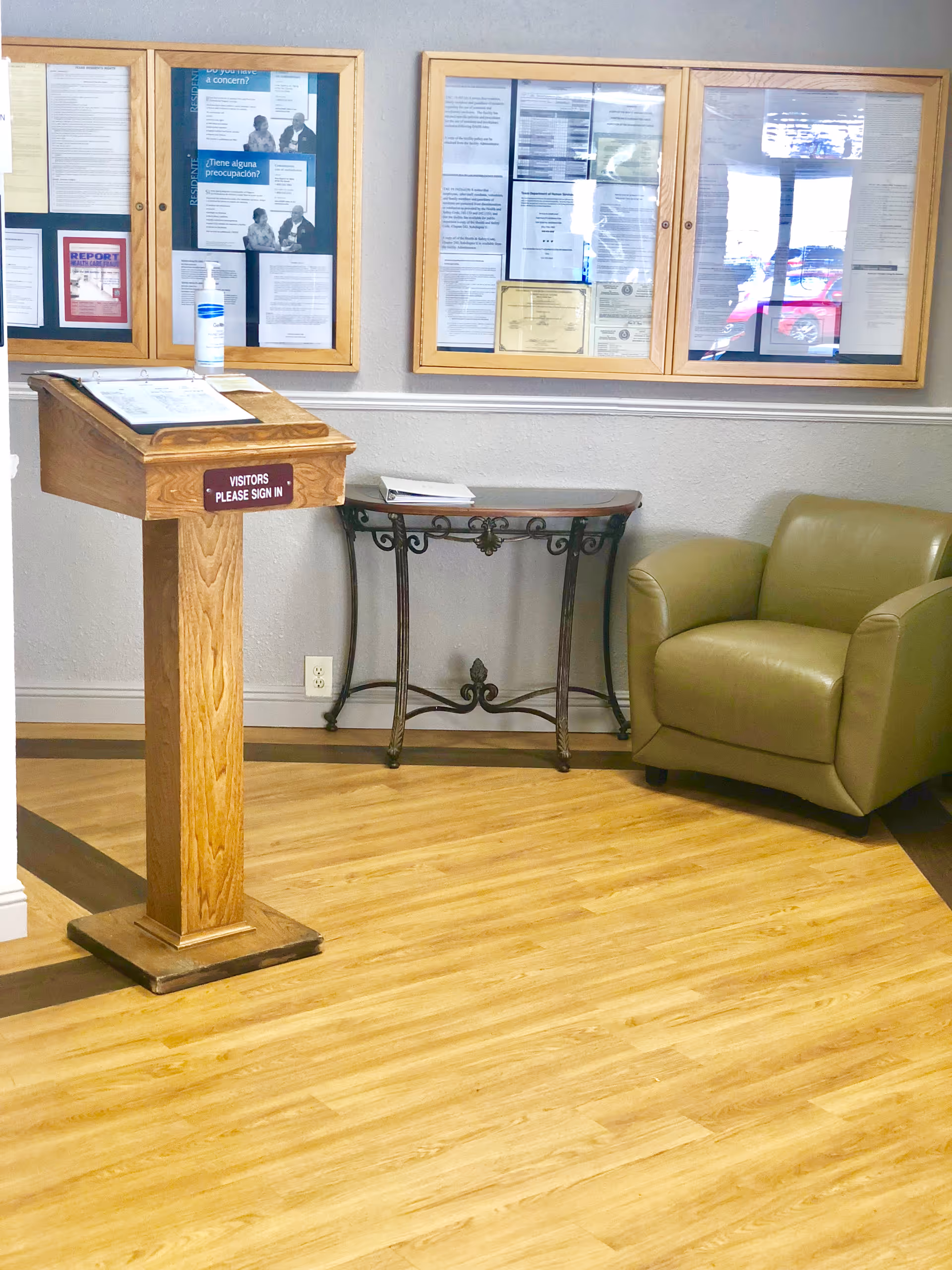 An interior corner of a facility with a wooden visitor sign-in stand, a small decorative table against the wall, a green armchair, and bulletin boards displaying various notices and documents on the wall.